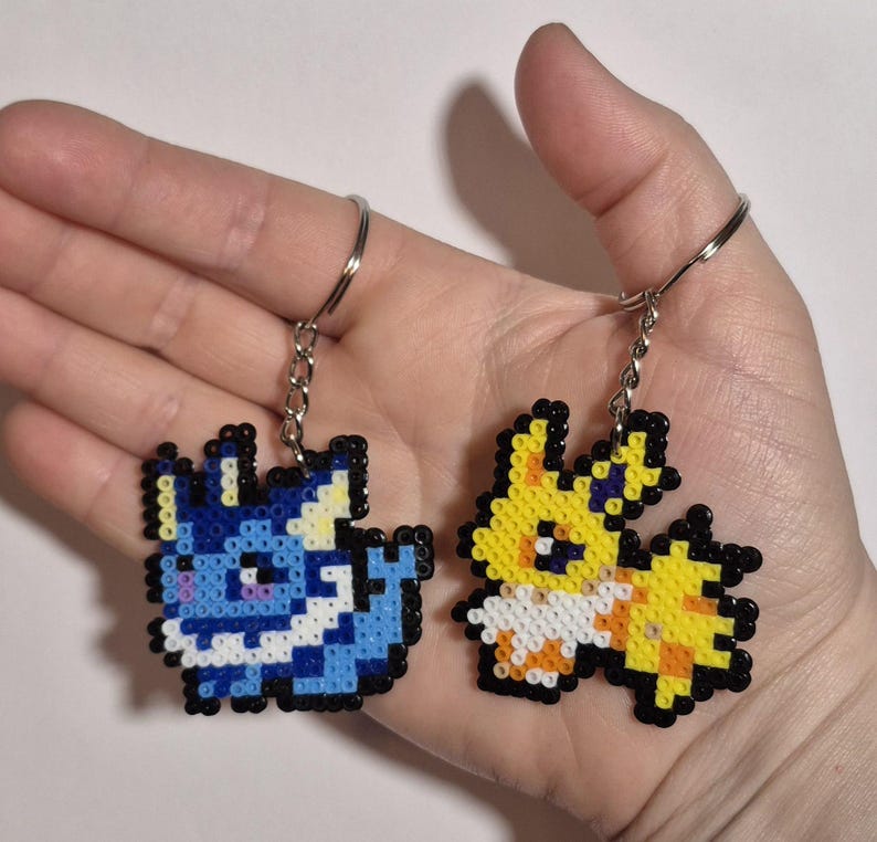 May include: Two pixel art keychains, one blue and one yellow, depicting cartoon characters. The blue keychain features a blue and white character with a white collar and yellow ears. The yellow keychain features a yellow and white character with a white collar and yellow ears.