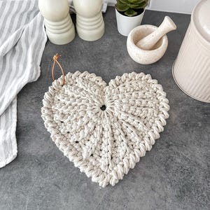 以下が含まれることがあります： クリーム色のハート型のマクラメ鍋敷き。吊り下げ用のループが付いています。厚手の織り綿ロープ製です。背景には、灰色のカウンタートップ、小さな鉢植えの多肉植物、キッチンアクセサリーが含まれています。