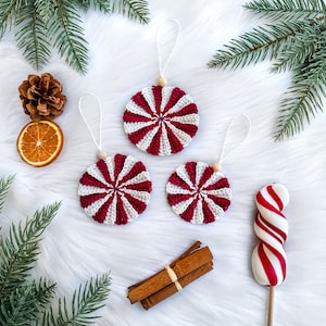 May include: Three red and white peppermint candy ornaments with a white string for hanging. The ornaments are on a white fluffy surface with a cinnamon stick bundle and a red and white candy cane.