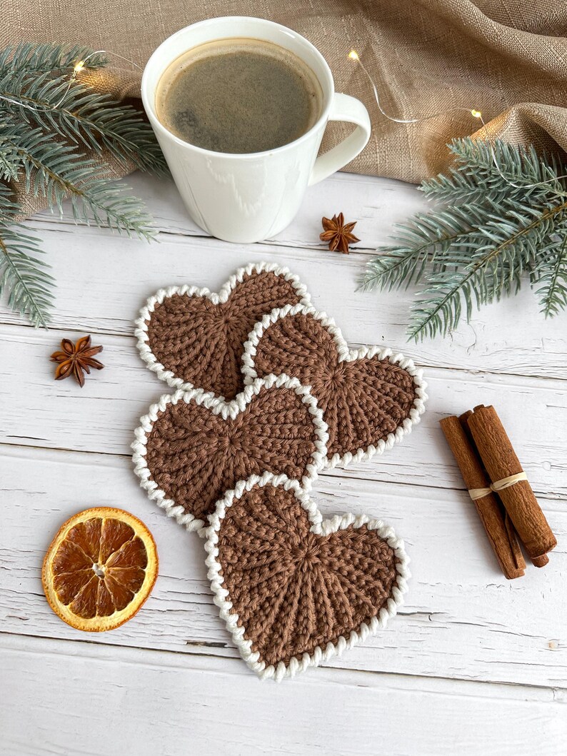May include: A white ceramic mug filled with coffee sits above four brown heart-shaped crochet coasters with white trim. The image also includes star anise, a dried orange slice, and cinnamon sticks, all arranged on a white wooden surface.