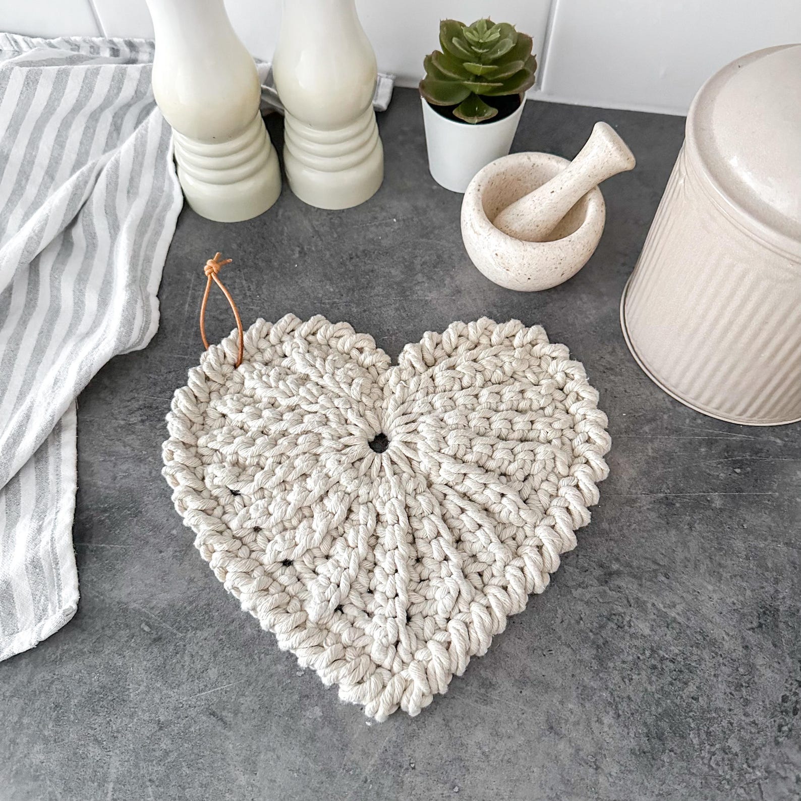 Crochet heart trivet or potholder in a chunky neutral yarn, styled on a gray kitchen counter with white salt and pepper shakers and a small plant.