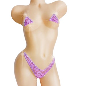 May include: A lavender bikini set with a triangle top and a thong bottom. The bikini is adorned with sparkling embellishments and delicate lace detailing. The set is displayed on a mannequin.