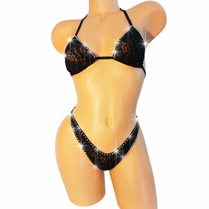 Pu&ograve; includere: Un set bikini in pizzo nero composto da un top a triangolo e uno slip sgambato. I bordi del bikini sono impreziositi da strass trasparenti e scintillanti che catturano la luce, creando un effetto luminoso sul tessuto in pizzo floreale.
