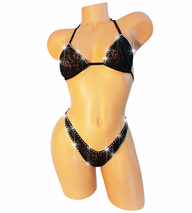 Pu&ograve; includere: Un set bikini in pizzo nero composto da un top a triangolo e uno slip sgambato. I bordi del bikini sono impreziositi da strass trasparenti e scintillanti che catturano la luce, creando un effetto luminoso sul tessuto in pizzo floreale.
