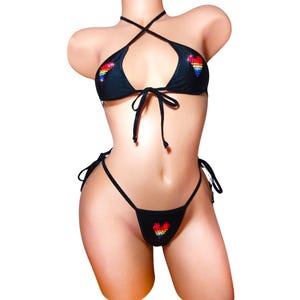 Puede incluir: Bikini negro con adornos de corazón arcoíris en las copas y la parte inferior. La parte superior del bikini tiene un estilo halter cruzado y se ata al cuello. La parte inferior es un bikini de hilo con lazos a los lados.