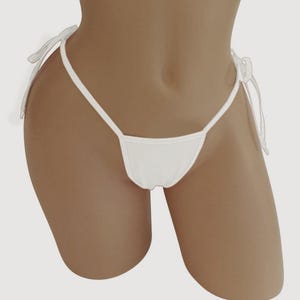 May include: White thong-style underwear with thin side straps that tie into bows. The front of the underwear has a triangular shape. The garment is made of a soft, opaque fabric.
