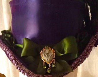 Può includere: Un cappello a cilindro viola con un nastro di raso verde e una spilla verde e dorata. Il cappello ha un bordo viola e bianco.
