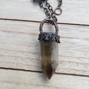 Puede incluir: Un collar con un colgante de cristal de cuarzo ahumado, una cadena de metal oscuro y un bisel de metal decorativo. El colgante es un cristal largo y puntiagudo de color marrón oscuro.