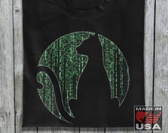 Op de afbeelding: Zwart T-shirt met een groen cirkelvormig ontwerp met een zwart katten silhouet. Het cirkelvormige ontwerp lijkt op de Matrix-code. Het shirt heeft de tekst "Made in USA" in de rechter benedenhoek.
