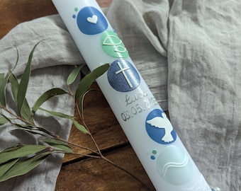 Pode incluir: Uma vela branca decorada com desenhos circulares azuis e verdes, incluindo um coração, uma cruz e uma pomba. A vela tem o nome "Luca" e a data "03.05.2023" impressos. Um detalhe de renda e fita adorna a base.