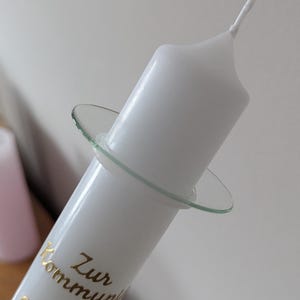Könnte beinhalten: Weiße Kerze mit einem Glas Halter. Die Kerze hat goldenen Text, der "Zur Kommunion" lautet. Die Kerze ist zylindrisch mit einem leicht ausgestellten oberen Ende und einem Docht.