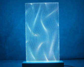 May include: A rectangular acrylic light panel featuring a series of vertical, undulating white lines that create a wave-like pattern. The panel is illuminated with a bright blue glow and is mounted on a solid, dark rectangular base with a power cord.