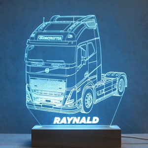 May include: An LED lamp featuring a clear acrylic panel etched with the outline of a Volvo Globetrotter semi-truck. The name RAYNALD is engraved in white at the bottom of the panel. The design is illuminated in light blue and sits on a wooden base.