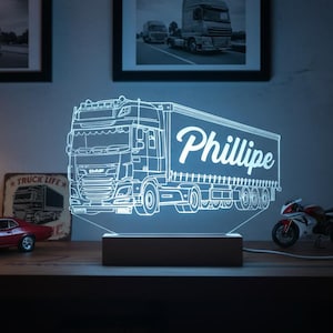 May include: A neon-lit truck-shaped lamp with the name "Phillipe" displayed on the trailer. The lamp sits on a wooden base, with a red car and motorcycle model nearby. Two framed truck photos hang in the background.