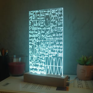 Puede incluir: Lámpara LED rectangular con fórmulas matemáticas grabadas. Emite una luz brillante y fría, sobre una base de madera. La lámpara está sobre una mesa con un libro, una vela y una pequeña planta en maceta. Dimensiones: aprox. 15 cm x 25 cm.