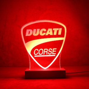 Könnte beinhalten: Ein rot beleuchtetes Ducati Corse Logo-Schild. Das Schild ist schildförmig und zeigt das Ducati-Logo oben und "CORSE" darunter. Es steht auf einem Holzsockel und leuchtet rot.