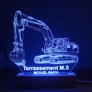 Può includere: Lampada LED blu a forma di escavatore con il testo "Terrassement M.S" e "Mickael Simon". La lampada presenta un contorno dettagliato di un escavatore su una base rettangolare, creando un'accattivante esposizione illuminata. Progettata per scrivania o tavolo.