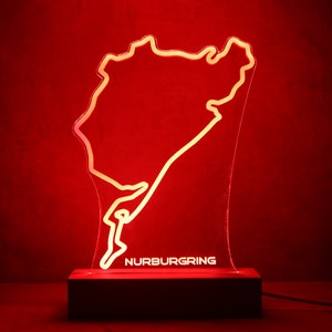 Puede incluir: Una pieza de exhibición de acrílico transparente que presenta el contorno rojo iluminado del circuito de carreras de Nürburgring. La palabra NURBURGRING está grabada en blanco en la parte inferior del panel acrílico, montado sobre una base rectangular.