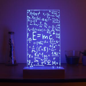 Op de afbeelding: Een blauw LED-licht met een wit acryl paneel waarop verschillende wetenschappelijke formules en vergelijkingen worden weergegeven, waaronder de beroemde vergelijking E=mc². Het licht staat op een witte basis en staat op een houten tafel.
