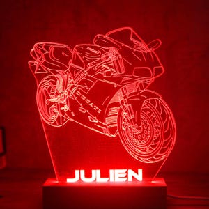 Może przedstawiać: Czerwona lampa LED z akrylu z szczegółową grafiką liniową motocykla Ducati. Podstawa lampy jest prostokątna i wyświetla imię JULIEN podświetlonymi białymi wielkimi literami na tle świecącego na czerwono wzoru motocykla.
