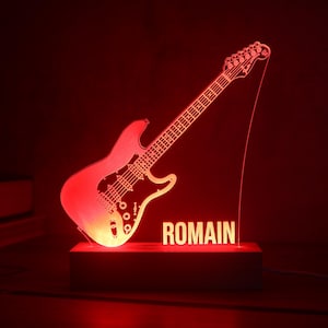 Può includere: Una lampada a LED rossa con una sagoma in acrilico trasparente di una chitarra elettrica e il nome ROMAIN in lettere maiuscole. Il design è montato su una base rettangolare in legno che illumina il pezzo in acrilico con un bagliore rosso.