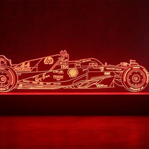 Puede incluir: Un contorno rojo iluminado de un coche de carreras de Fórmula 1, con logotipos de patrocinadores y el número 44. La luz está montada sobre una base negra, creando un efecto visual llamativo. El diseño del coche se destaca con un brillo brillante y cálido.
