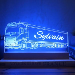 Peut inclure: Une lampe LED en acrylique personnalisée présentant une illustration au trait détaillée d'un semi-remorque Volvo avec une remorque. Le prénom Sylvain est gravé en écriture cursive sur le côté de la remorque. La lampe émet une lumière bleue vive.