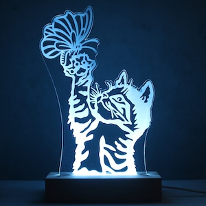 Puede incluir: Lámpara LED con forma de gato y diseño de mariposa. La lámpara está hecha de acrílico transparente y se asienta sobre una base de madera. El gato extiende la pata para tocar la mariposa. La lámpara emite un suave brillo azul.