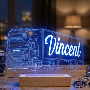 Peut inclure: Une lampe LED en acrylique transparent présentant le contour gravé d'un semi-remorque Scania avec le nom Vincent écrit en lettres cursives sur la remorque. La lampe est fixée sur un socle en bois rectangulaire et émet une lumière bleue.