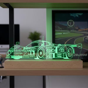 Op de afbeelding: Een groene LED-lamp met de omtrek van een Mazda 787B racewagen, voorzien van het nummer 55 en de teksten RENOWN en CHARGE aan de zijkant. Het acryl silhouet van de auto is gemonteerd op een rechthoekige, lichtgekleurde houten voet.