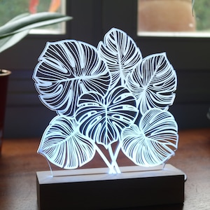 Op de afbeelding: Een witte, verlichte, acryl lamp met een ontwerp van vijf tropische bladeren. De lamp staat op een houten voet en staat op een houten tafel.