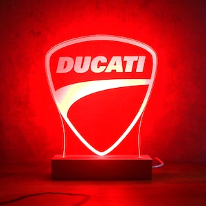 Könnte beinhalten: Rotes Ducati-Logo-Nachtlicht. Das Ducati-Schildlogo ist weiß mit weißem Markennamen. Das Licht steht auf einem rechteckigen Holzsockel und leuchtet hell vor rotem Hintergrund.