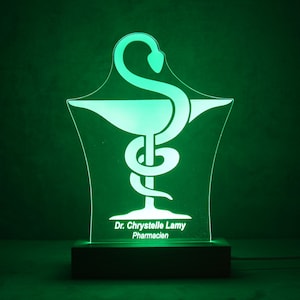 Puede incluir: Una luz de noche verde iluminada con el símbolo de la farmacia. La luz presenta el símbolo de la vara de Asclepio y el texto "Dr. Chrystelle Lamy Pharmacien" sobre una base negra. La luz está enchufada y brillando.
