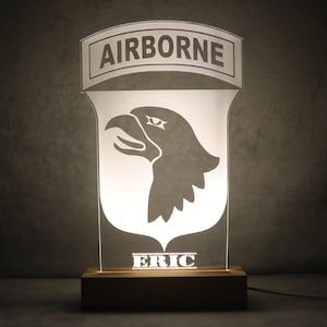 Op de afbeelding: Verlichte acryllamp met het woord "AIRBORNE" boven een silhouet van een adelaarskop. De naam "ERIC" wordt onderaan weergegeven. De lamp is gemonteerd op een houten voet en heeft een netsnoer.