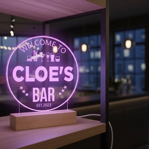 Peut inclure: Enseigne lumineuse LED rose pour "Cloe's Bar". Le panneau rond affiche "Welcome to", des graphiques de bar et "EST.2023". Il est monté sur une base en bois, idéal pour un bar.