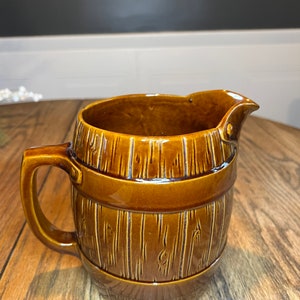 Pode incluir: Um jarro de cerâmica marrom em forma de barril com um design de grão de madeira. O jarro tem uma alça e um bico.