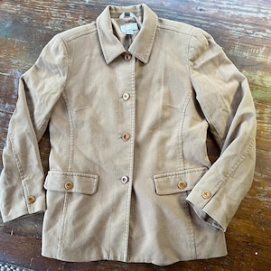 May include: A tan-coloured blazer featuring a structured collar, long sleeves with buttoned cuffs, and a front button closure. The garment includes two front flap pockets and a visible brand label at the inner neckline, showcasing a classic design.