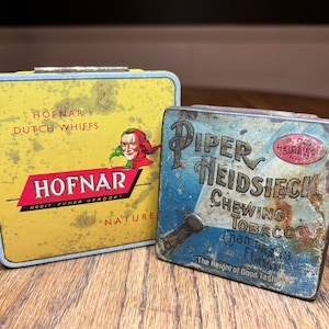Puede incluir: Dos latas de metal vintage para tabaco. La lata de la izquierda es amarilla con letras rojas y negras que dicen "HOFNAR DUTCH WHIFFS HOFNAR NOOIT FUNER GEROORT NATURE". La lata de la derecha es azul y blanca con letras rojas y doradas que dicen "PIPER HEIDSIECK CHEWING TOBACCO Champagne Flavor The Height of Good Tast".
