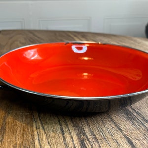 May include: A black enamel bowl with a red interior and a white rim. The bowl has two black handles on the sides.