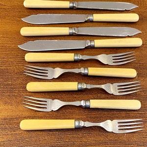 May include: A set of vintage cutlery, comprising knives and forks, displayed on a wooden table. The knives feature silver blades and cream-coloured handles. The forks have silver tines and matching cream handles. This silverware set is likely for use at a dining table.