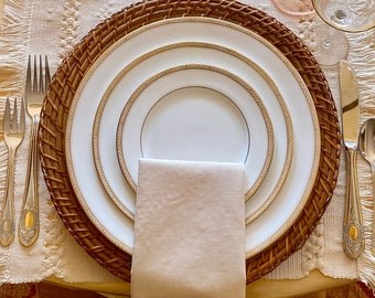 May include: A set of white ceramic dinner plates with gold trim, nested inside a woven wicker charger plate. A beige linen napkin is placed in the center of the plates.