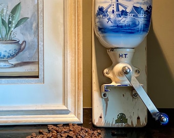 May include: A vintage blue and white ceramic coffee grinder with a windmill design. The grinder is mounted on a wooden base and has a metal crank. A pile of coffee beans are in the foreground.