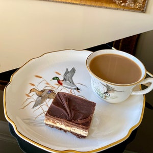 May include: A white porcelain plate with a gold rim features a brown and white chocolate dessert with a chocolate topping. A cup of coffee and a small plate with a bird design are also on the plate.