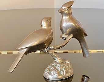 May include: A brass sculpture of two birds perched on a branch. The birds are stylized and have a smooth, metallic finish. The sculpture is on a black surface.