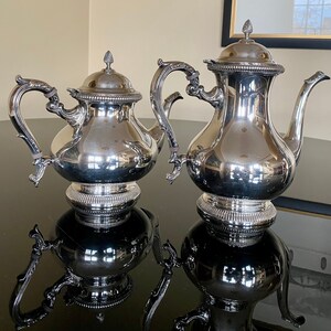 May include: Two silver teapots with ornate handles and spouts. The teapots are polished and reflect the surrounding environment.