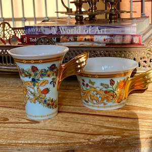 May include: Two white ceramic teacups with gold winged handles and floral patterns. The cups are on a wooden surface.