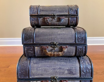 May include: Three vintage suitcases stacked on top of each other. The suitcases are made of woven material and have a dark brown color. The top suitcase has a brass latch and a brown leather handle. The middle suitcase has a brass latch and a brown wooden handle. The bottom suitcase has a brass latch and a brown leather handle.