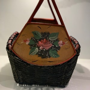 May include: A black wicker basket with a red painted handle and a painted floral design on the top. The floral design features a pink rose with green leaves and black berries.