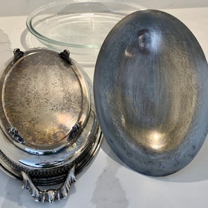 Op de afbeelding: Een ovale serveerschaal van zilver met een decoratieve rand en sierlijke handvatten. De schaal heeft een bijpassend deksel met een glad, licht gewelfd oppervlak.