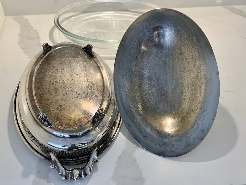 Op de afbeelding: Een ovale serveerschaal van zilver met een decoratieve rand en sierlijke handvatten. De schaal heeft een bijpassend deksel met een glad, licht gewelfd oppervlak.